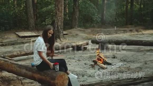 一个女孩坐在森林里看书。 女孩坐在火旁边的一根木头上，看一本书，特写。