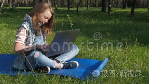 一个女孩坐在公园里拿着笔记本电脑。 一个十几岁的女孩坐在地毯上呼吸新鲜空气。 阳光明媚的一天，一个带着笔记本电脑的女孩
