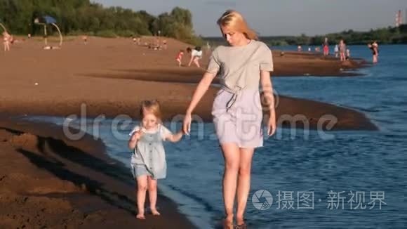 美丽的金发妈妈和女儿在日落时在海滩上散步。