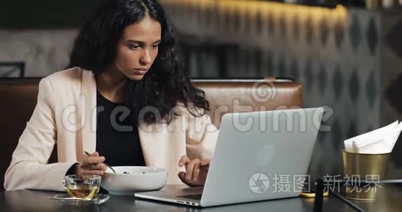 商务女性坐在咖啡馆里，拿着笔记本电脑吃晚饭，桌上放着一杯茶。 上班吃午饭
