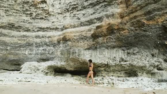 在海滩上穿泳衣的女孩。 在巨大石壁的背景下.. 她沿着海滩散步。 她走路