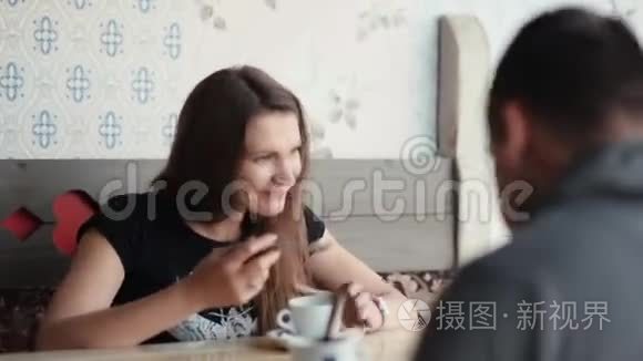 漂亮的黑发女人和男人在咖啡馆里。 她舔勺子，享受咖啡或茶。 她说话，微笑，玩得很开心。