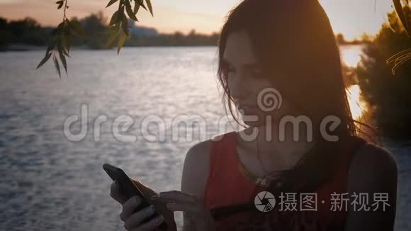 迷人的年轻女性在日落时使用智能手机。 一个可爱的女孩微笑着在海上使用手机。