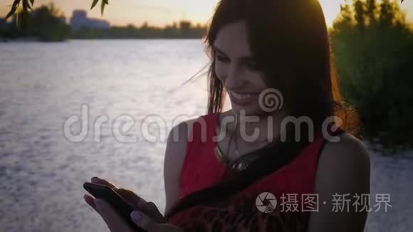 迷人的年轻女性在日落时使用智能手机。 一个可爱的女孩微笑着在海上使用手机。