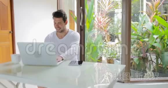 年轻人在家工作，手拿电脑，坐在桌子上，微笑着打字