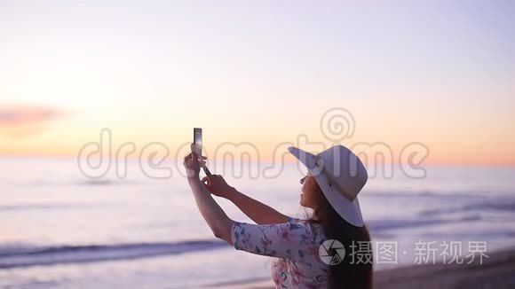 戴帽子的年轻女游客在海洋海滩日落时拍照