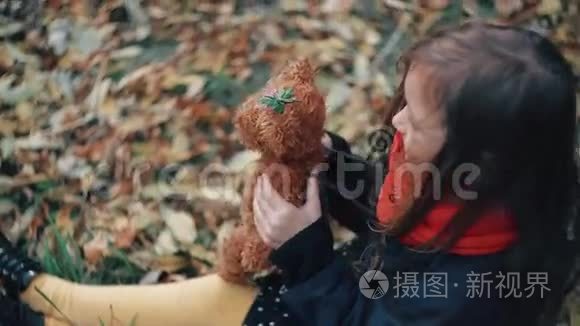 可爱的小女孩玩她的泰迪熊拥抱，并把它抱在她坐在草地上的秋天公园4k。