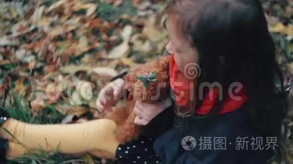 可爱的小女孩玩她的泰迪熊拥抱，并把它抱在她坐在草地上的秋天公园4k。