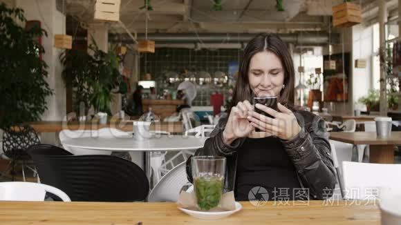 时尚的年轻女士喝着草药茶，用智能手机坐在现代咖啡馆的桌子上，微笑着。 4K