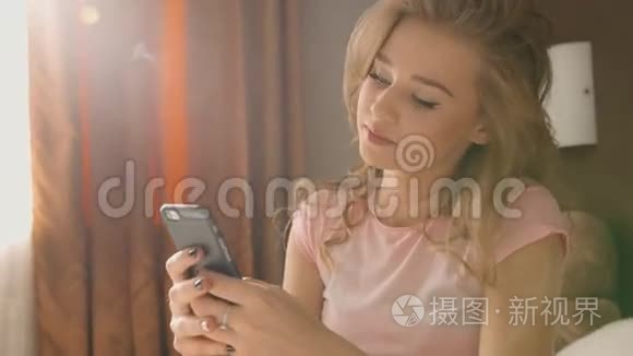 金发女人坐在床上，手里拿着手机，聊天，听到朋友穿着粉红色的睡衣。