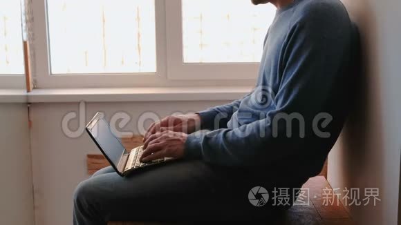 在笔记本电脑上工作。 一个面目全非的年轻人坐在靠近窗户的阳台上，在笔记本电脑里打字。