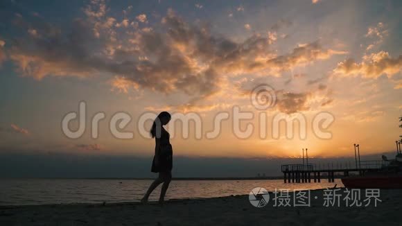 日落时海滩上一个穿着浅色连衣裙的女孩的剪影。 美丽的少女赤脚走在沙滩上。 慢动作