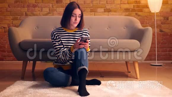 在室内一间舒适的公寓里，一个年轻漂亮女孩用手机坐在地板上的特写镜头