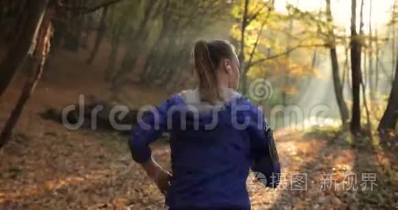 公园里的女跑步者。 穿着蓝色夹克和其他健身服的漂亮女人跑步时听音乐