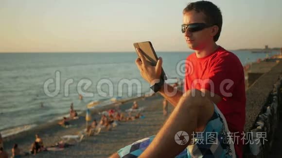 年轻的帅哥戴着太阳镜，带着平板电脑在海边放松。 他查看社交网络上的信息
