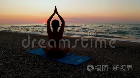 美丽的年轻女子坐在海滩上做瑜伽，一边看日落。