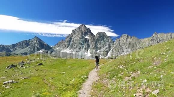 女人在田园诗般的山景中跋涉，在小径上穿越盛开的绿色草地，坐落在高高的岩石山上