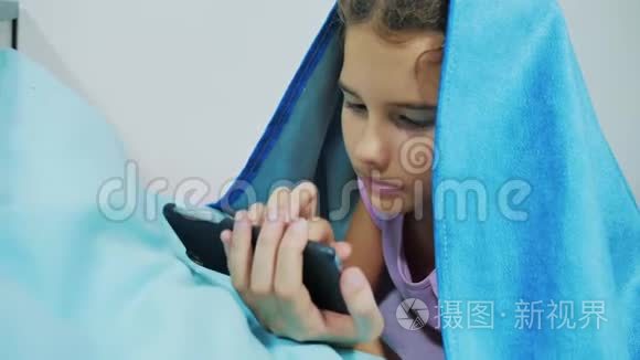 带智能手机的毯子下可爱女孩的肖像。 在社交生活方式媒体下玩网络游戏的小女孩