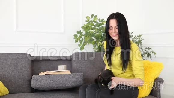 对宠物的爱。 美丽的黑发女孩带着一只黑猫在家里的沙发上客厅里放松。