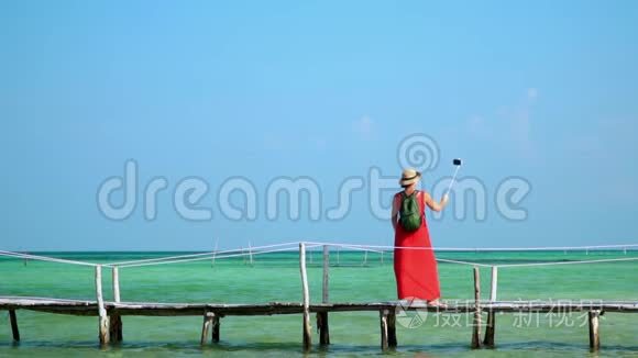 穿红色衣服的女人带着绿色背包在码头上散步。 旅行者把自己的照片贴在赛尔夫棍子上