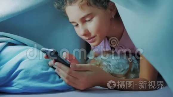 可爱的女孩和狗宠物的肖像在毛毯下与智能手机。 小女孩和狗宠物在网上玩游戏