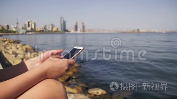 年轻女性使用智能手机坐在海边的堤岸和梅格波利斯摩天大楼上