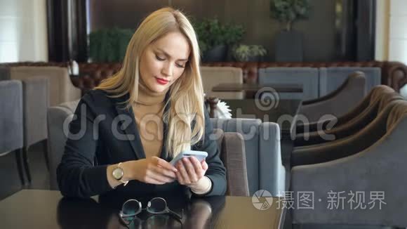一个金发商务女性，穿着西装，拿着电话坐在咖啡馆的桌子旁