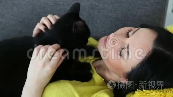 对宠物的爱。 美丽的黑发女孩带着一只黑猫在家里的沙发上客厅里放松。