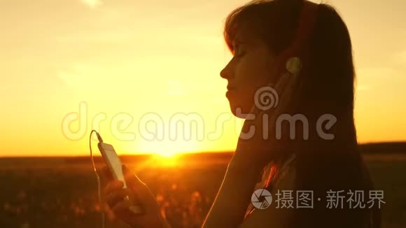 快乐的女孩听音乐，在美丽的夕阳下跳舞。 戴着耳机和智能手机的年轻女孩