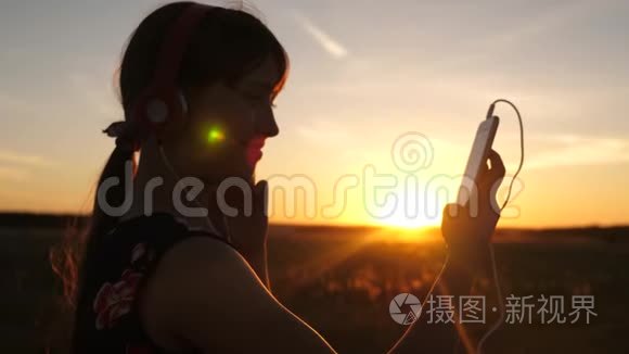 快乐的女孩在美丽的夕阳下，在智能手机上听音乐。 戴着耳机的年轻女孩