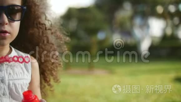 快乐的孩子在公园里吹肥皂泡。 漂亮的卷发小女孩戴着红眼镜吹着肥皂泡