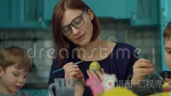 30岁的年轻母亲和两个儿子在家里的蓝色厨房里画复活节彩蛋。 准备复活节的幸福家庭