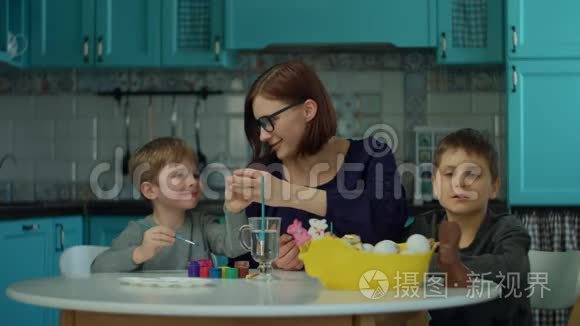 30岁的年轻母亲和两个儿子在家里的蓝色厨房里画复活节彩蛋。 准备复活节的幸福家庭