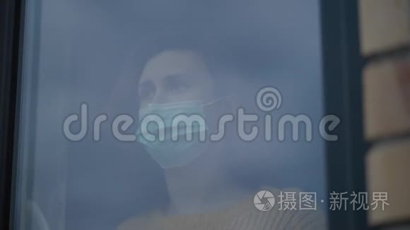 戴着外科口罩的窗后年轻悲伤的女人，在自我隔离概念中，冠状病毒流行时看得很远