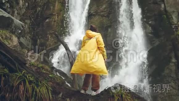 穿着黄色雨衣的年轻男性旅行者喜欢美丽的瀑布。 在山里远足。 徒步旅行者跑到