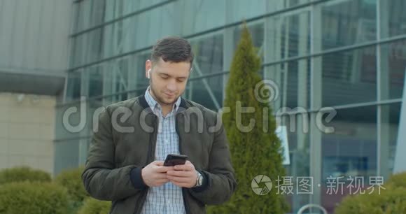 带智能手机和耳机的人在现代办公中心附近行走的肖像。 在社交网络上和他聊天