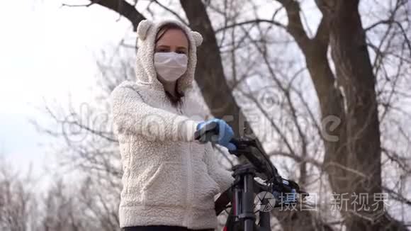 戴着医用口罩和手套的年轻女子骑着自行车站在农村，手握舵。 保护妇女