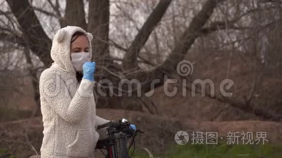 戴着医用口罩和手套的年轻女子咳嗽着，手握着农村自行车的舵。 保护妇女