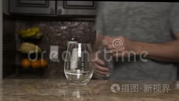 穿着灰色T恤的年轻人在一个漂亮时尚的厨房里用热水瓶或塑料瓶把水倒入玻璃杯中