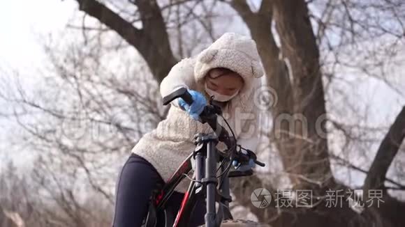 戴着医用口罩和手套的年轻女子骑着自行车站在农村，手握舵。 保护妇女