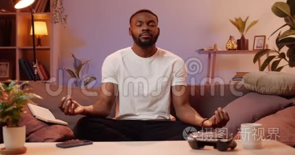 年轻的帅哥在家里坐在沙发上做呼吸锻炼。 在放松的时候做瑜伽的非洲男人