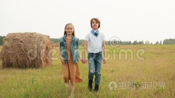 十几岁的夫妇在干草堆背景下漫步在农村的田野上。 快乐的青少年女孩和男孩牵着手继续走