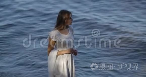 悲伤的黑发白种人女人穿着浅色的白色连衣裙站在海浪的背景上思考。 有魅力的女孩