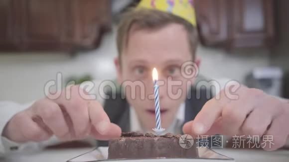 一个戴着生日帽的滑稽年轻人坐在厨房里的一个小巧克力蛋糕前面的肖像。 书呆子有