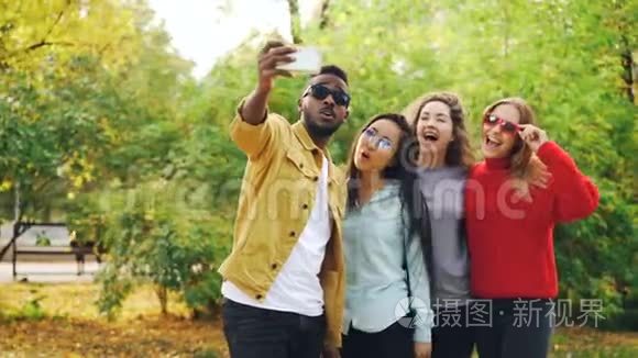 非洲裔美国学生正在与美丽的女孩自拍，亚洲和高加索人站在公园里，使用智能手机