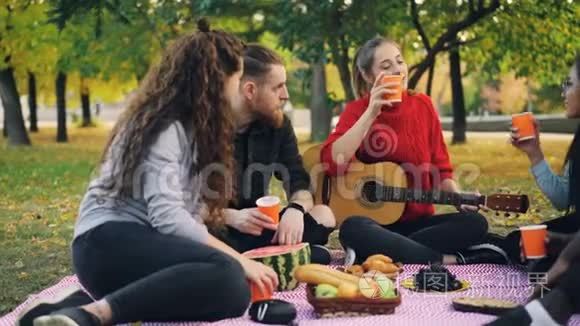 在温暖的秋日，年轻的男人和女人在公园野餐时用吉他品尝和敲击眼镜。 友谊