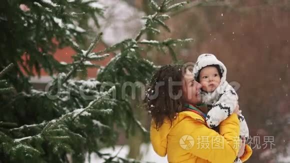 可爱的年轻女子穿着黄色羽绒服和婴儿穿着工作服走路。 下雪了，妈妈给他的儿子看，