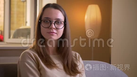 一位年轻迷人的白种人女性戴着眼镜，看着坐在室内沙发上的摄像机。