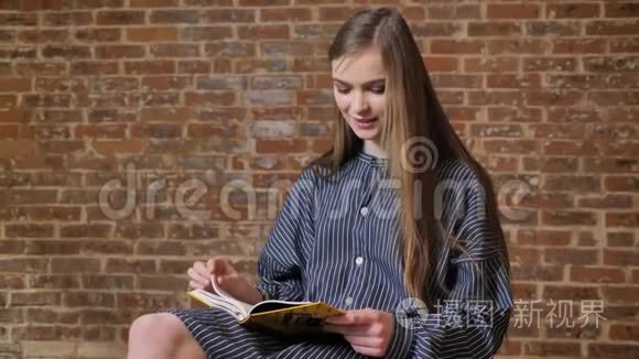 年轻漂亮的女孩坐着看书，看着相机，微笑着，放松的概念，砖背景